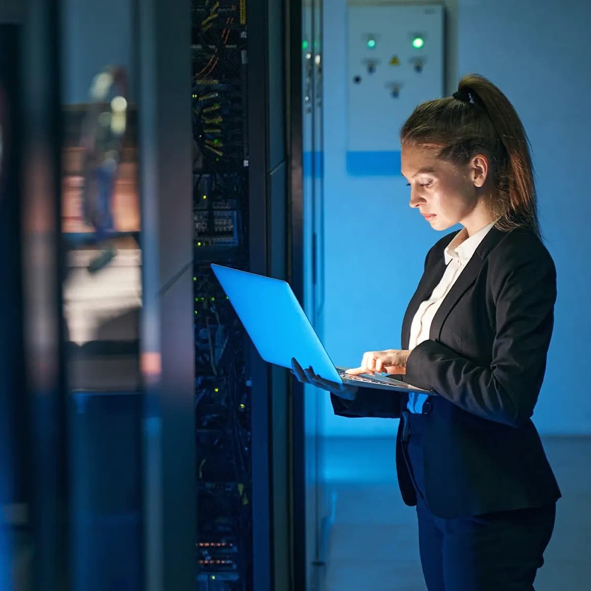 Woman with a laptop in front of a data center rack enclosure.